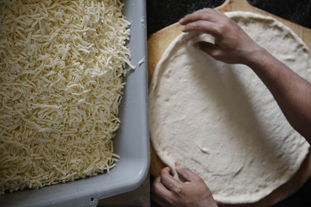 Pizza maker stretching dough next to a bin of shredded mozzarella cheese